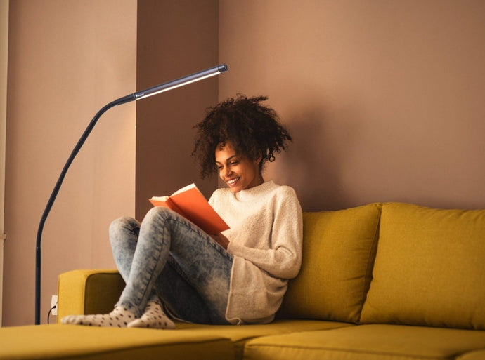 Lady sitting on mustard couch using Electra lamp to read an orange book. 